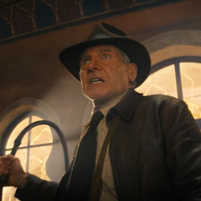 En arg Indiana Jones (Harrison Ford) med läderrock och hatt står med piskan i handen i en stor och hög sal.