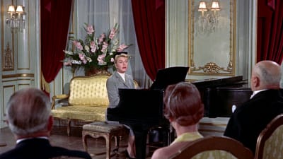 Nainen (näyttelijä Doris Day) soittaa ja laulaa flyygelin ääressä hienosti sisustetussa salongissa, etualalla selin istuvia kuulijoita. Kuva elokuvasta Mies joka tiesi liikaa.