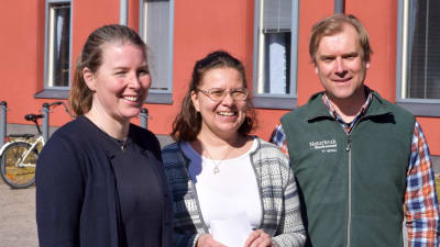 Två kvinnor och en man utomhus utanför en skolbyggnad.