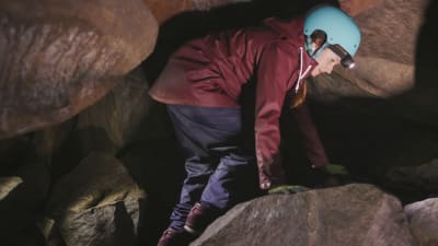 Kvinna i blå hjälm och pannlampa går hukad mellan stenar inuti en grotta.