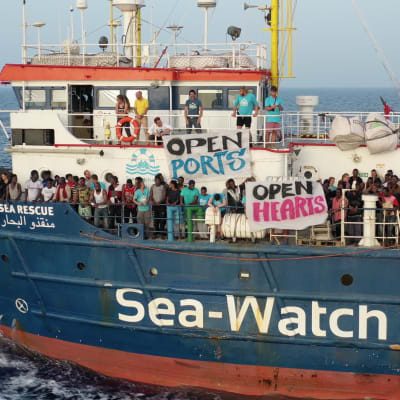 En bild på båten Sea Watch med flyktingar ute på däcket med skyltar där det står "open ports" och "open hearts".