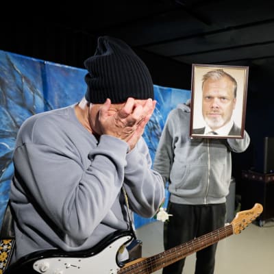 En man med en elgitarr kring axlarna döljer ansiktet i händerna. En annan håller upp ett inramat porträttfoto framför sitt ansikte.