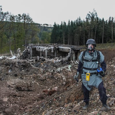 Suojapukuun, kaasunaamariin ja kypärään pukeutunut mies seisoo räjähtäneen näköisessä maisemassa tuhoutuneen betonisen rakennelman edessä.Taustalla näkyy metsämaisemaa.