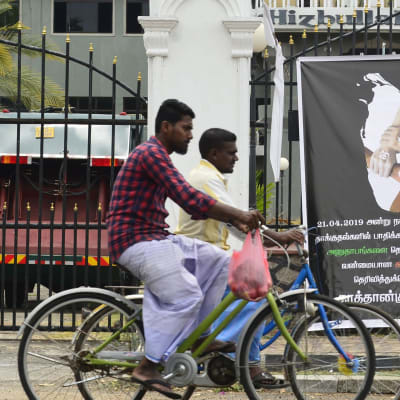 Cyklister på Sri Lanka vid en plansch som visar fyra händer med olika religiösa symboler