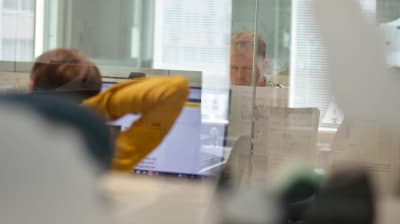 En kvinna sträcker på sig. Hon sitter bakom en glasdörr och jobbar vid en dator. I kontorslandskapet sitter också en man.
