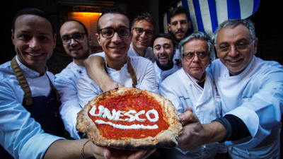 Åtta pizzabagare håller upp en napolitansk pizza där det står skrivet Unesco.