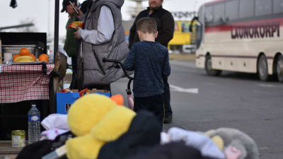 Liten pojke och några vuxna ute på en parkeringsplats vid Ukrainska gränsen i Polen. En låda med leksaker i förgrunden och en buss i bakgrunden.