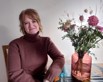 Fyrtiofemårig kvinna sitter intill en blomvas på sitt matbord. Hon ler in i kameran.