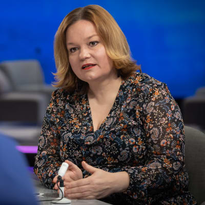 Krista Kiuru sitter vid ett bord och talar. I förgrunden syns en redaktörs huvud bakifrån.