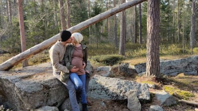 En man och en gravid kvinna kramas och pussas i skogen. De lutar sig mot en sten.