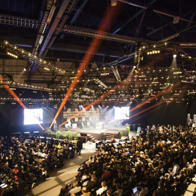 Massor med människor sitter, står och följer med konferensen Slush 2014 i en stor arena med färggranna strålkastare.