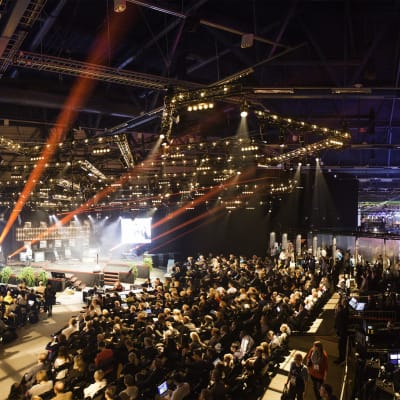 Massor med människor sitter, står och följer med konferensen Slush 2014 i en stor arena med färggranna strålkastare.
