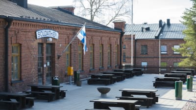 Låg röd tegelbyggnad med texten soldathem, utanför vajar Finlands flagga. Det finns bänkar och bord utanför.