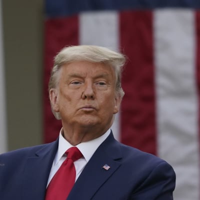 Man i blå kostym och röd slipps står framför amerikansk flagga. Mannen är USA:s president Donald Trump.