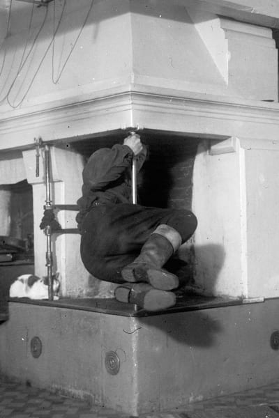 En man leker leken "gå omkring spisjärnet", där han hänger i järnstören i en öppen spis. Bilden är tagen 1937 i Korsnäs, Harrström.