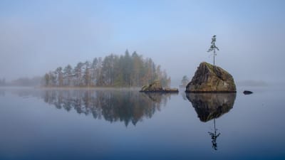 Kuvassa on Suomessa utuiseen järven pintaan heijastunut metsäinen saari ja iso kivi.