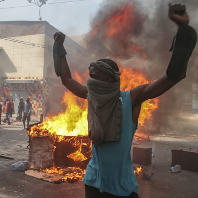 Mielenosoittaja seisoo kädet ylhäällä palavien esineitten edessä Dakarissa.