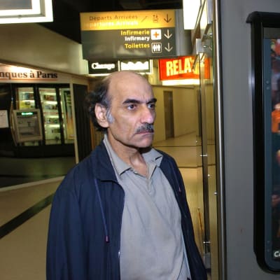 En man med buskiga ögonbryn och mustasch står bredvid en reklampelare med den franska filmaffischen för Steven Spielbergs film The Terminal.