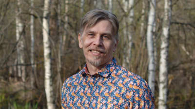 Porträtt av Harri Myllyniemi.Han poserar med en spräcklig skjorta och ser in i kameran. I bakgrunden syns björkar. . 