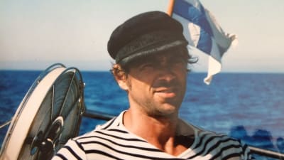En man står på en segelbåt i randig skjorta och seglarmössa. I bakgrunden finlands flagga och blått hav.
