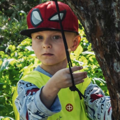 Liten pojke i gul reflexväst och röd skärmmössa står i en lummig trädgård och leker med ett rep som hänger från ett äppelträd.