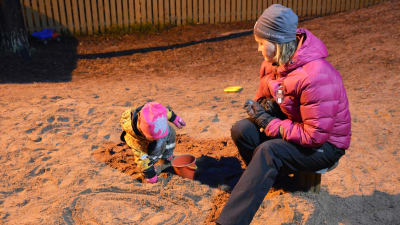 Ett litet barn leker i en sandlåda. Bredvid barnet sitter en kvinna i rosa jacka och ser på. Det är mörkt ute men sandlådan lyses upp av gårdens lampor.
