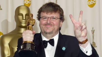 Dokumentaristi, kirjailija Michael Moore nostaa Oscar-patsasta ja näyttää voitonmerkkiä, takanaan suuri Oscar-patsas.