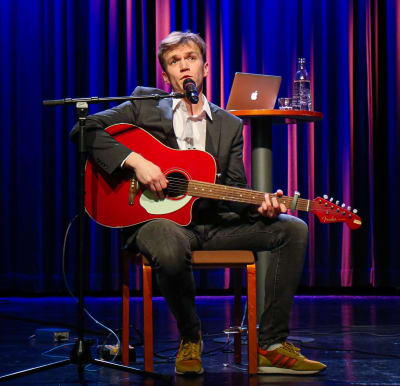 Alfred Backa sitter på en stol på en scen framför ett draperi. Han har en gitarr i famnen och pratar in i en mikrofon.