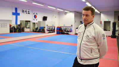 En man i vit skjorta står inne i en idrottshall. På väggen finns speglar samt Finlands och Koreas flagga. Golvet är rödblå gummimatta.
