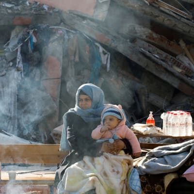 Ett litet barn i sin mammas famn, i bakgrunden syns ruiner efter jordbävning.