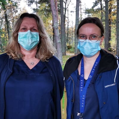 Koronakartoittajat Gun Grangärd ja Sari Kauppinen seisovat katoksen alla hoitajavaatteissaan ja maskit kasvoillaan.