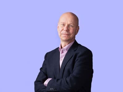 En skallig leende man i mörk kostym mot en lila bakgrund.