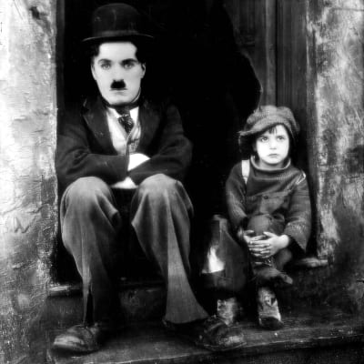 Knallipäinen kulkuri (Charlie Chaplin) ja pieni poika (Jackie Coogan) istuvat rappusilla. Kuva elokuvasta Chaplinin poika.
