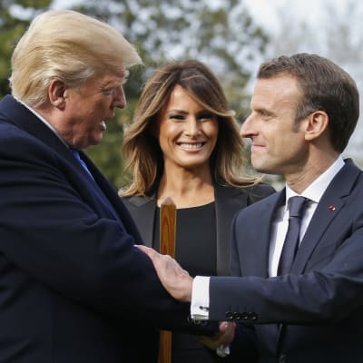Presidenterna Emmanuel Macron och Donald Trump skakar hand medan Melania Trump ser på och ler.