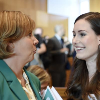 Anna-Maja Henriksson och Sanna marin talar med varandra. I bakgrunden syns Li Andersson, i förgrunden en suddig Katri Kulmuni.