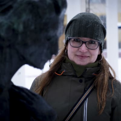 Riikka Miettinen katsoo kameraan. Etualalla näkyy naispatsaan kasvojen ääriviivat.