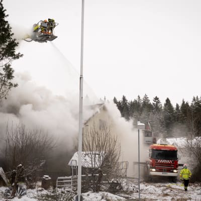 Lumisella pihalla paloauto, ilmassa nostolavalla palomiehiä suihkuttamassa vettä runsaasti savuavan talon katolle.