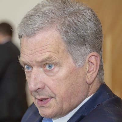 I förgrunden syns en suddig axel, i mitten av bilden sitter Sauli Niinistö.
