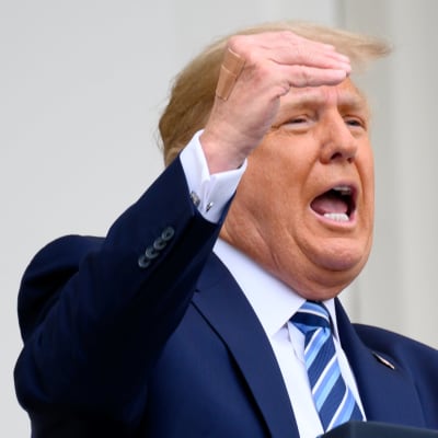 Trump puhuu parvekkeella mikrofoniin nostaen oikeaa kättään. Hänellä on tummansininen puku ja vinoraidallinen kravatti.