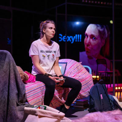 Näyttelijä Jutta Järvinen istuu sängyllä puhelin kädessä ja katselee kaukaisuuteen näytelmässä Usko, toivo, huijaus