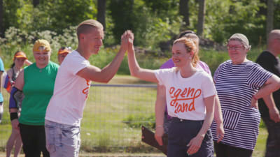 Mies ja nainen löyvät kädet yhteen hymyillen kesällä urheilukentällä, taustalla hymyileviä ihmisiä.