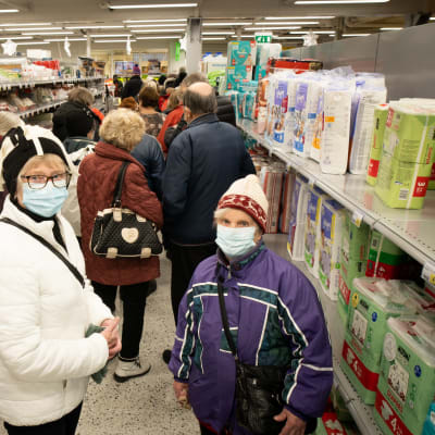 Kaksi talvivaatteisiin ja kasvomaskiin pukeutunutta naista katsoo kameraan kaupan käytävällä. Hyllyillä myytäviä tavaroita, naisten edessä paljon ihmisiä jonossa.
