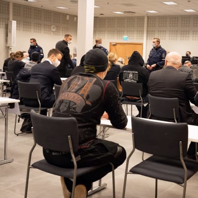 Åtalade sitter med ryggen till, i bakgrunden poliser.