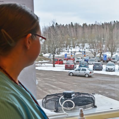 Kvinna i sjukskötardräkt tittar ut genom ett fönster mot en parkeringsplats.