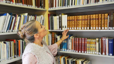 En blond kvinna står vid en bibliotekshylla fylld med böcker. Hon håller handen på en bokrygg och tittar uppåt mot böckerna ovanom henne.