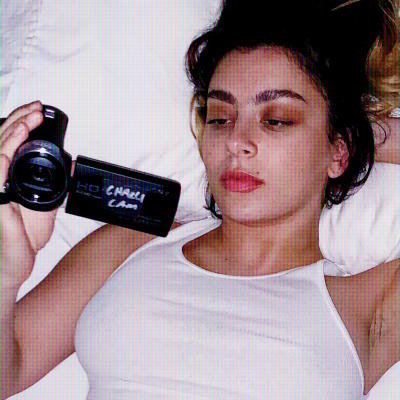 Charli XCX makaa alusvaatteissaan sängyllä ja kuvaa itseään pikkukameralla.