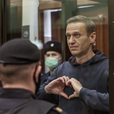 Aleksej Navalnyj står i en glasbur omgiven av poliser och formar ett hjärta med händerna.   