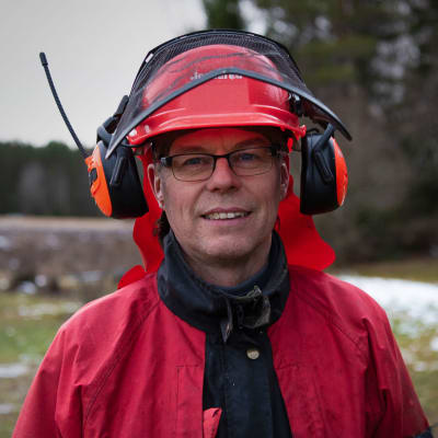 närbild av en man med glasögon, röd rock och en skogsarbetares skyddshjälm med tillhörande hörselskydd