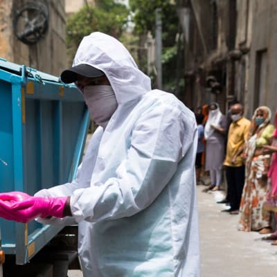 En kommunalt anställd i New Delhi får sina handskar desinficerade efter att han tagit hand om sopor i en lokal där man erbjuder coronatester. I bakgrunden står folk och köar för att testas. 