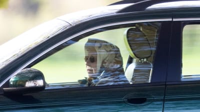 Drottning Elizabeth kör bil. Hon har solglasögon på sig och en skarf runt huvudet. Bilden är taget genom fönstret på förarsidan av bilen.